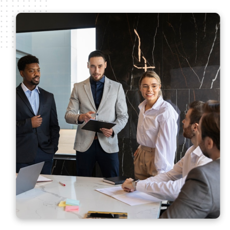 Five people in business attire are having a discussion around a conference table in a modern office, collaborating on strategies for the best CRM software. One stands holding a clipboard, while others listen and smile amid sticky notes and papers.