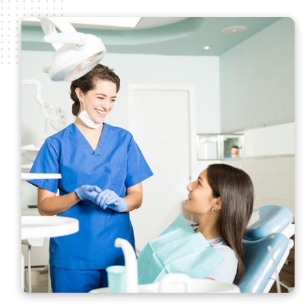 A dentist in blue scrubs and gloves smiles while talking to a patient sitting in a dental chair, in a bright and modern dental office powered by the best CRM software for seamless patient management.