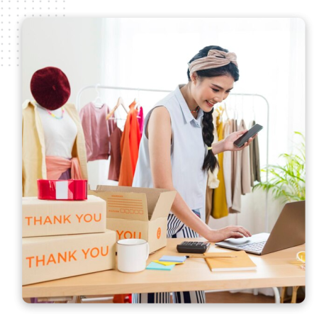 A woman stands at a desk with a laptop, smiling while holding a phone. Around her are thank you boxes, tape, a coffee mug, and clothing on a rack, suggesting she uses the best CRM software to run her online shop from home.