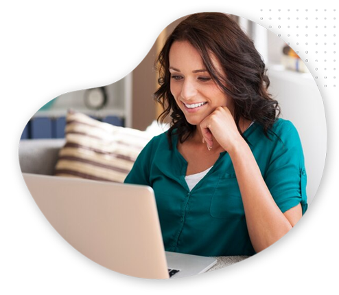 A woman with dark hair, wearing a teal blouse, sits at a table and smiles while looking at a laptop screen in a bright, cozy room with striped pillows in the background.
