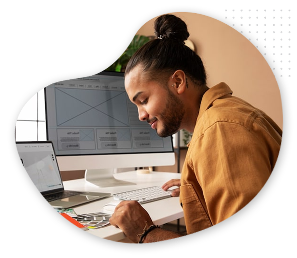 Blog-web-img A person with long hair in a bun, wearing a brown shirt, sits at a desk working on web design wireframes on the computer, with a laptop nearby, holding a pen and color swatches—perfect inspiration for your next blog post.