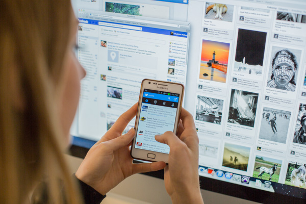 A person holding a smartphone and browsing social media, with a computer monitor displaying multiple social media feeds in the background—perfectly illustrating an effective Social Media Strategy.