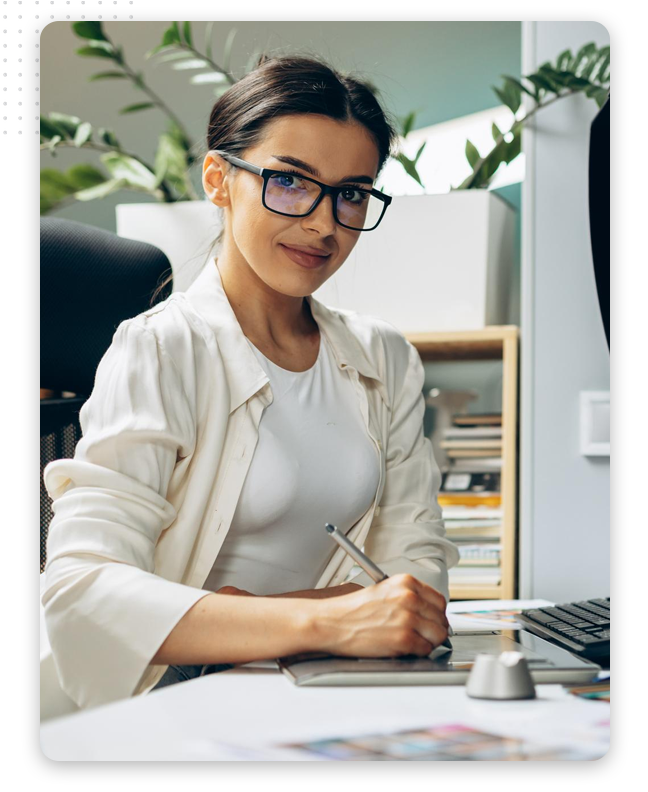 A woman wearing glasses and a white shirt sits at a desk, smiling and holding a stylus pen, working on paperwork and a graphic tablet—surrounded by plants and shelves, she appears focused on digital design projects.
