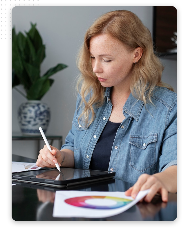 logo-design-process-feat-img A woman with long blonde hair wearing a denim shirt uses a stylus on a tablet while holding a color wheel printout at a desk, embodying the creative process of professional logo designers, with a potted plant in the background.