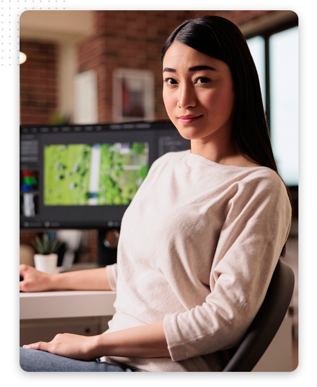 video-production-process-feature-img A woman with long dark hair sits at a desk, looking at the camera and smiling slightly. Behind her, a computer screen displays a digital map in the modern office of a video production company.
